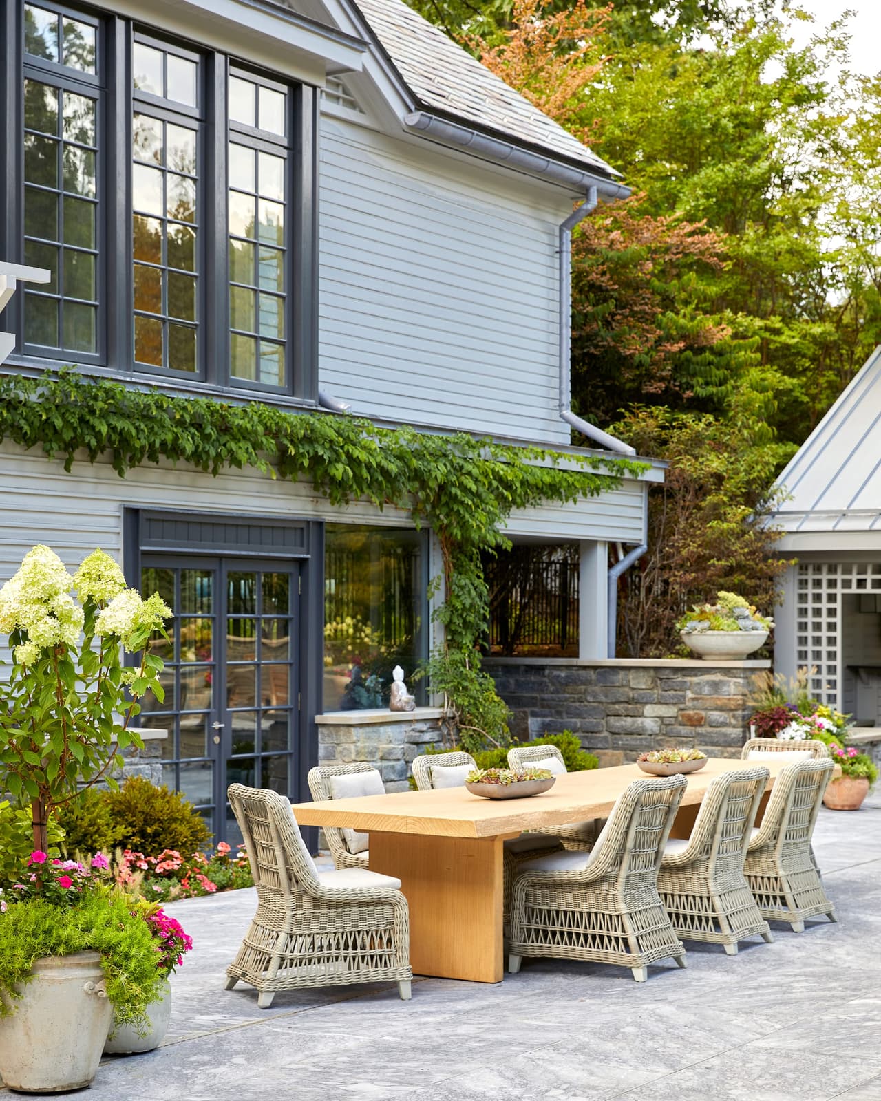 Long outdoor dining table and wicker chairs on the terrace of a Westfield, NJ home, against blue-gray shingles with climbing vines and potted hydrangeas