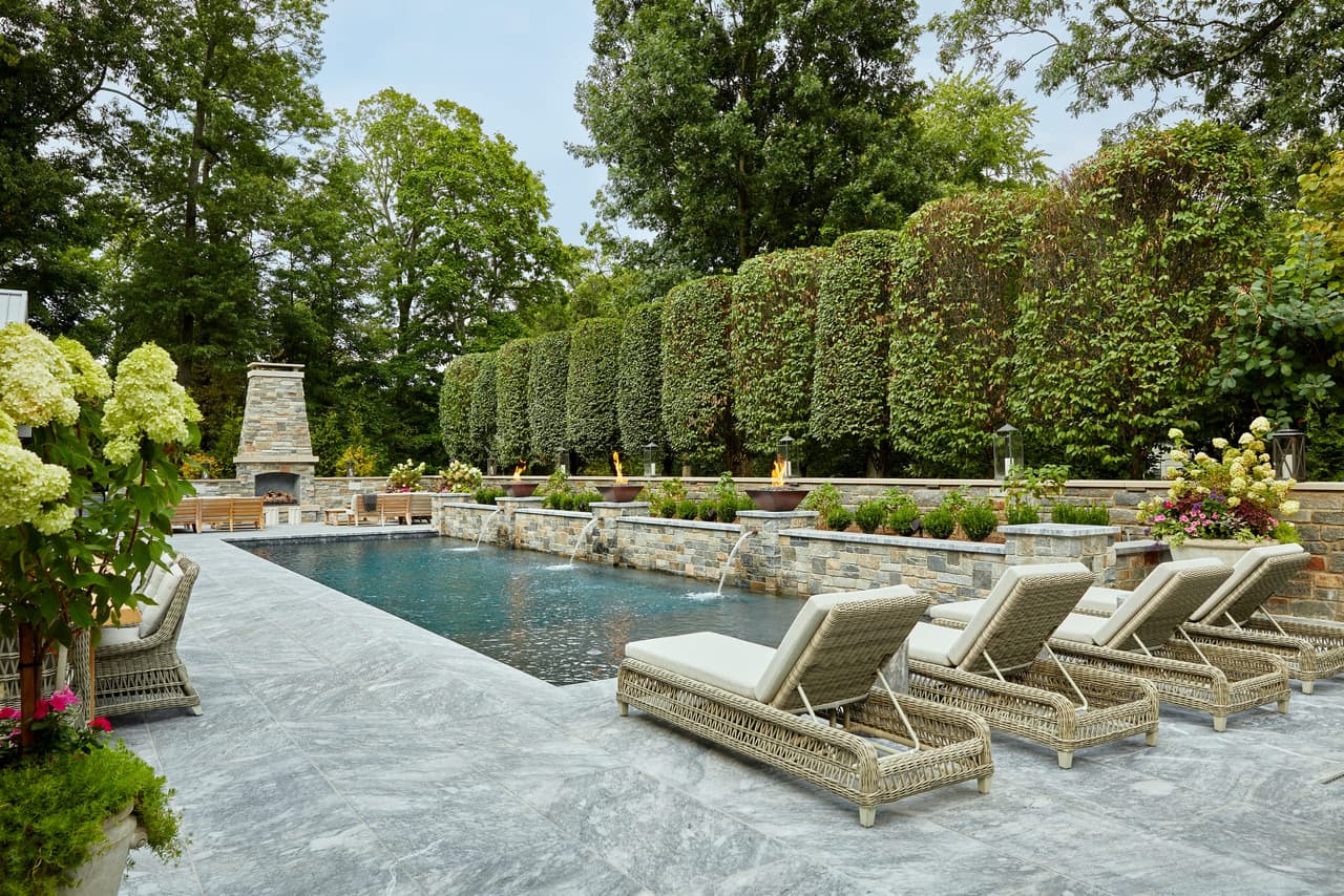 Pool and chaise seating on a bluestone terrace behind a Westfield, NJ home, with a clipped hedge along the boundary and a stone outdoor fireplace