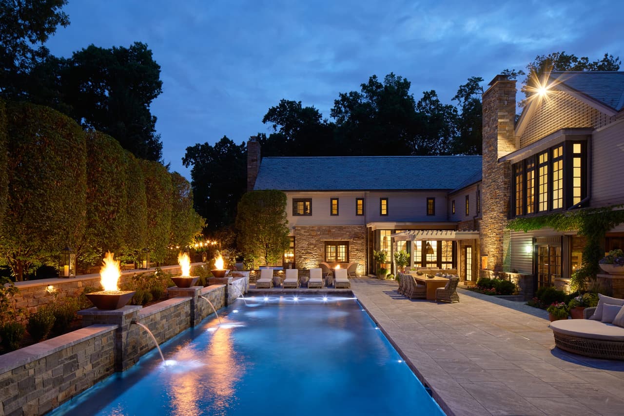 Long evening view down the lit pool of a Westfield, NJ backyard, with fire bowls burning along the terrace and the house lit behind