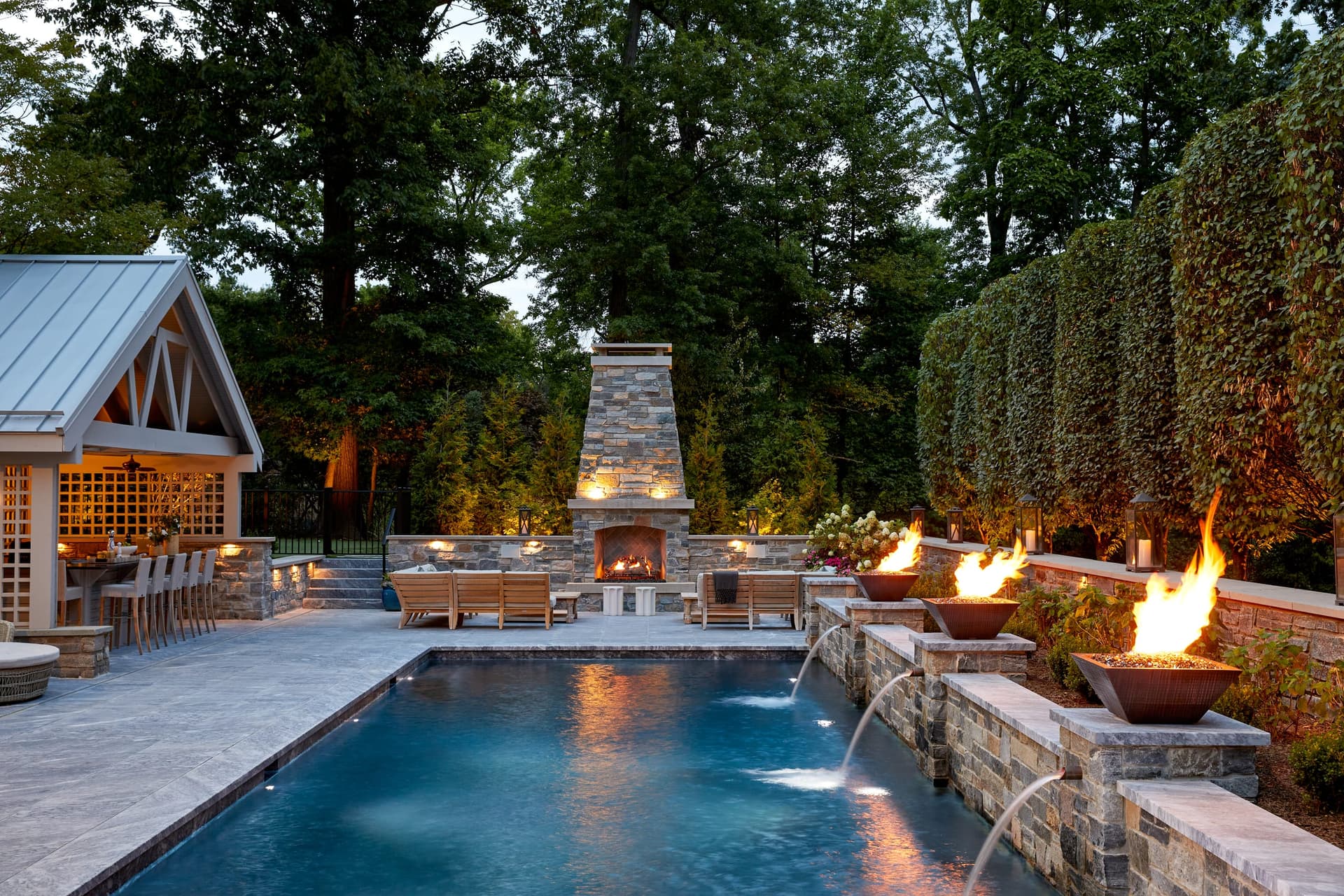 Westfield, NJ backyard at dusk with the pool lit, fire bowls alight along the raised stone wall, an outdoor fireplace and a cedar pergola beyond, built by Michael Robert Construction