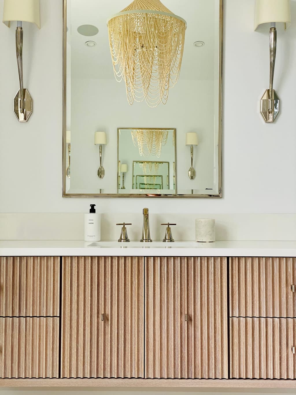 Fluted oak vanity under a brass-framed mirror between two sconces in the primary bathroom of a Westfield, NJ new-construction home, with a beaded chandelier above