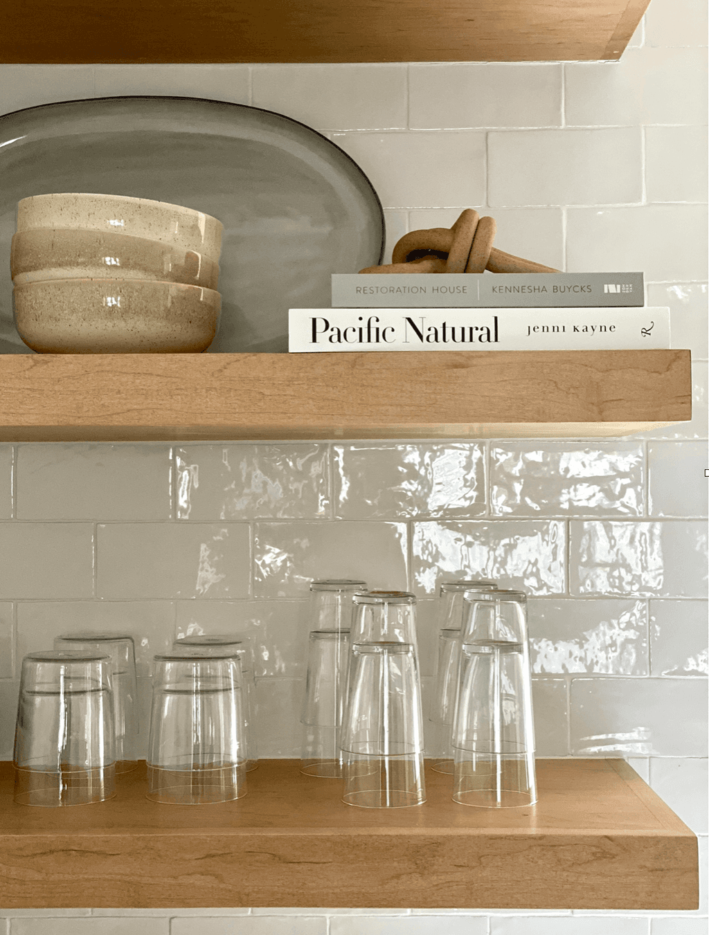 Open oak shelves above a glazed tile backsplash, holding a cookbook, ceramics and glassware