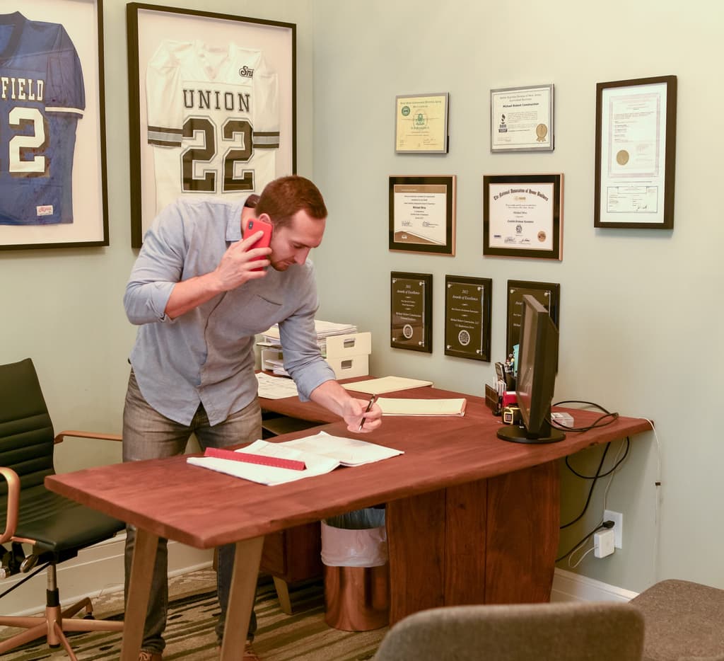 Mike Mroz on the phone at his desk in the Westfield, NJ office, beneath framed awards and jerseys