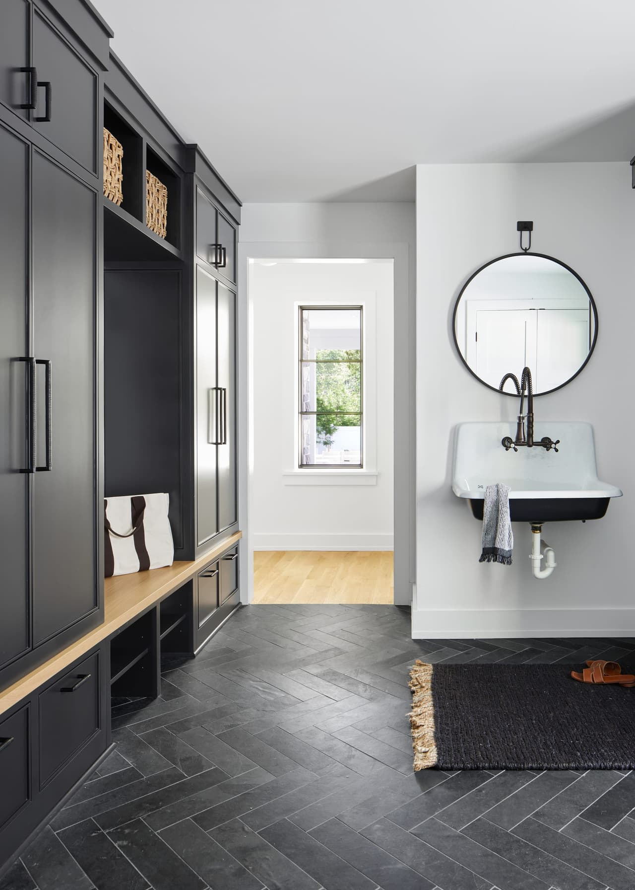 Mudroom of a Westfield, NJ home renovation with charcoal custom built-ins, an oak bench, a wall-hung sink and a herringbone stone floor