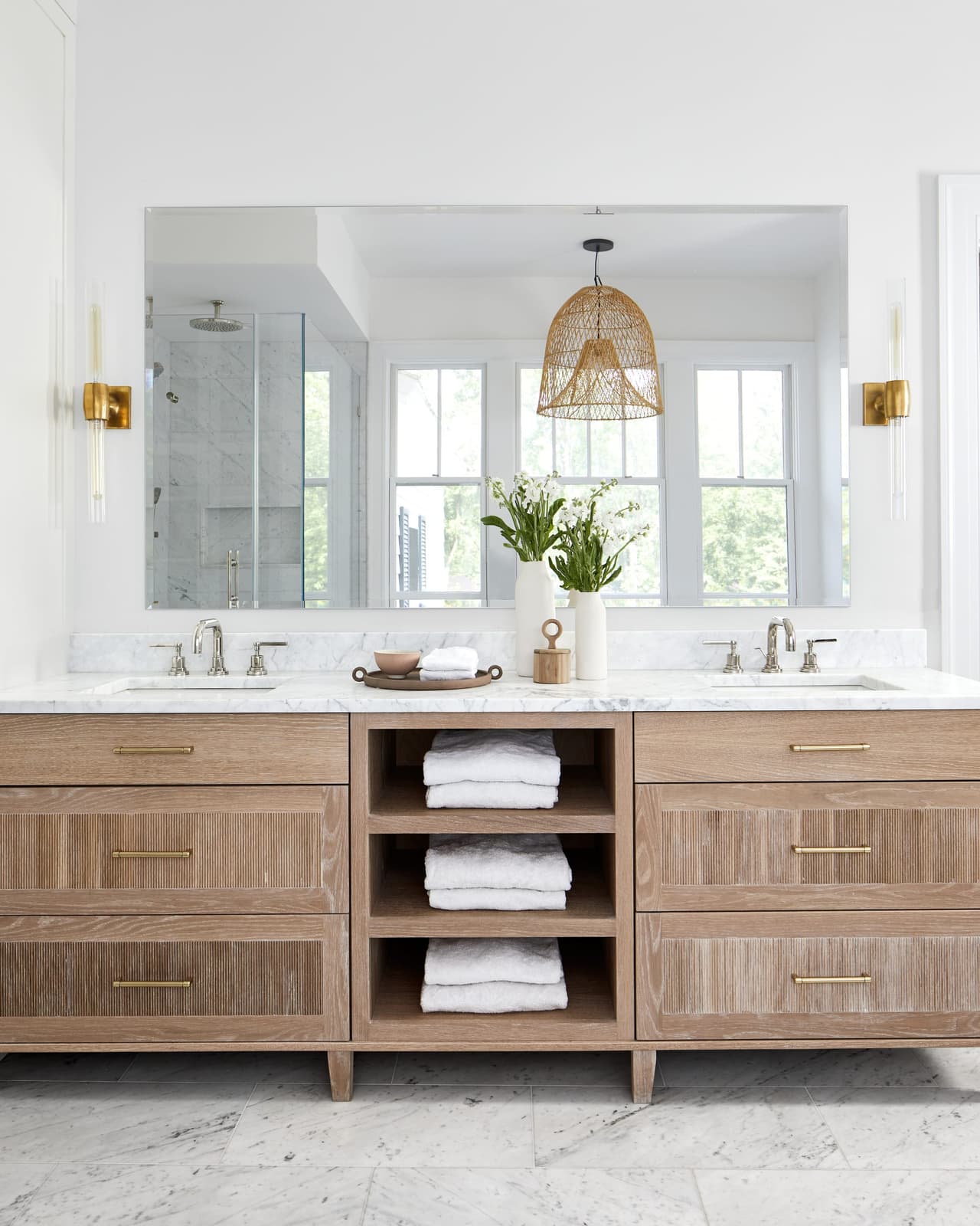 Primary bathroom in Westfield, NJ with a natural wood double vanity, open towel shelves, a white marble top and a woven pendant over the walk-in shower