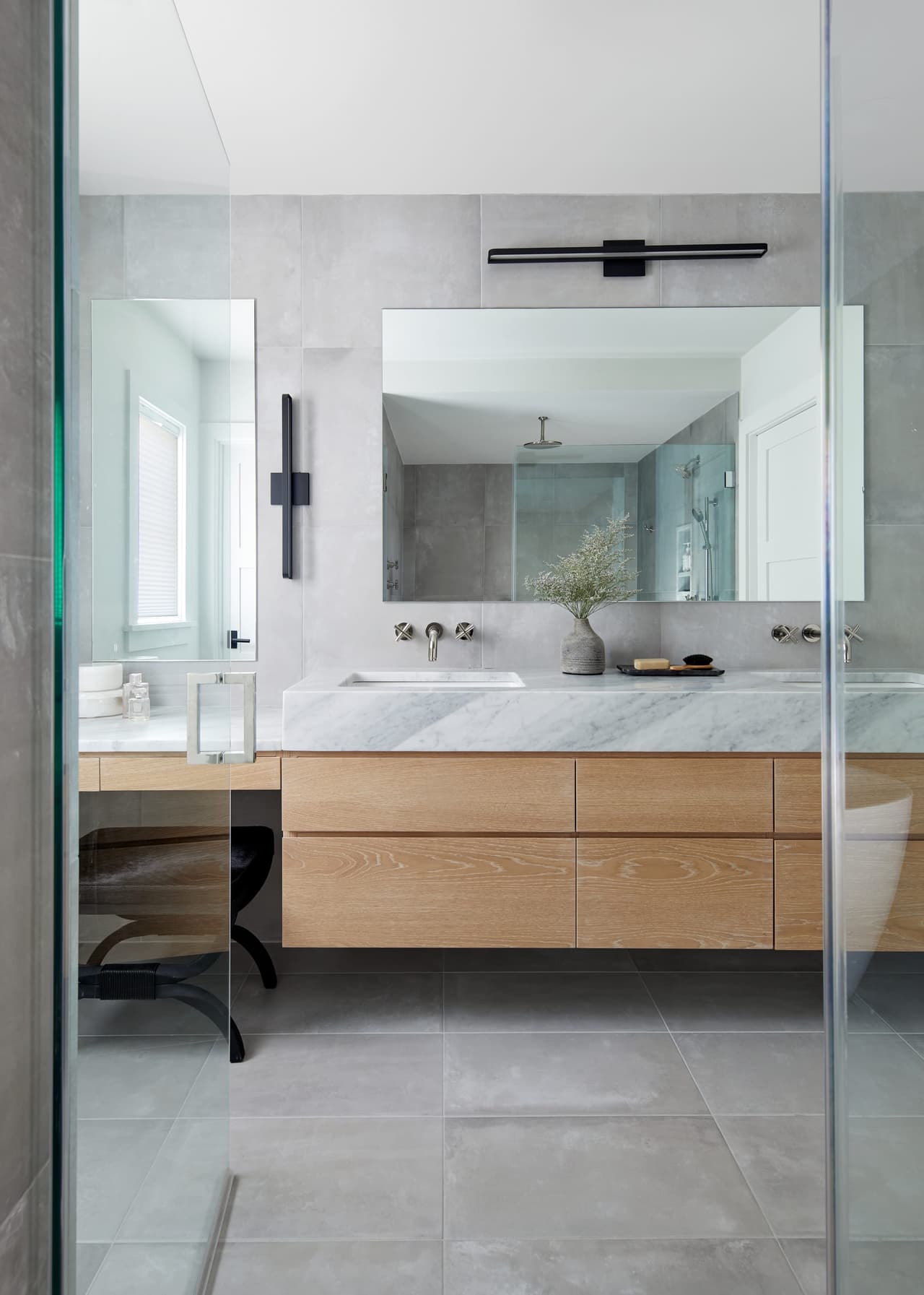 Primary bathroom of a Westfield, NJ home with a floating white oak vanity, a marble top and a long mirror lit from above, seen past the glass shower