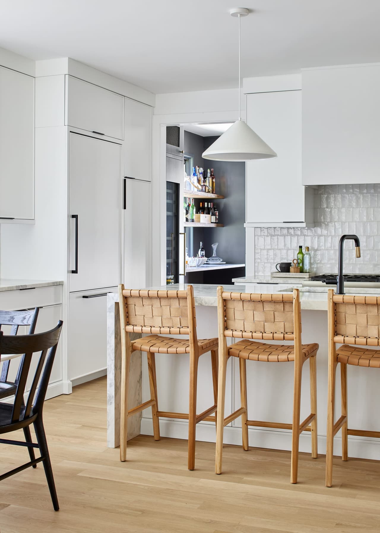 White modern kitchen in a Westfield, NJ home renovation, with woven leather counter stools, black hardware and a glass-front bar cabinet beyond