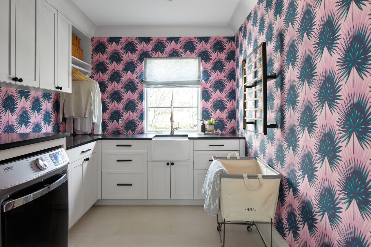 Laundry room with bold pink and teal palm wallpaper, white cabinetry and a side-by-side washer and dryer