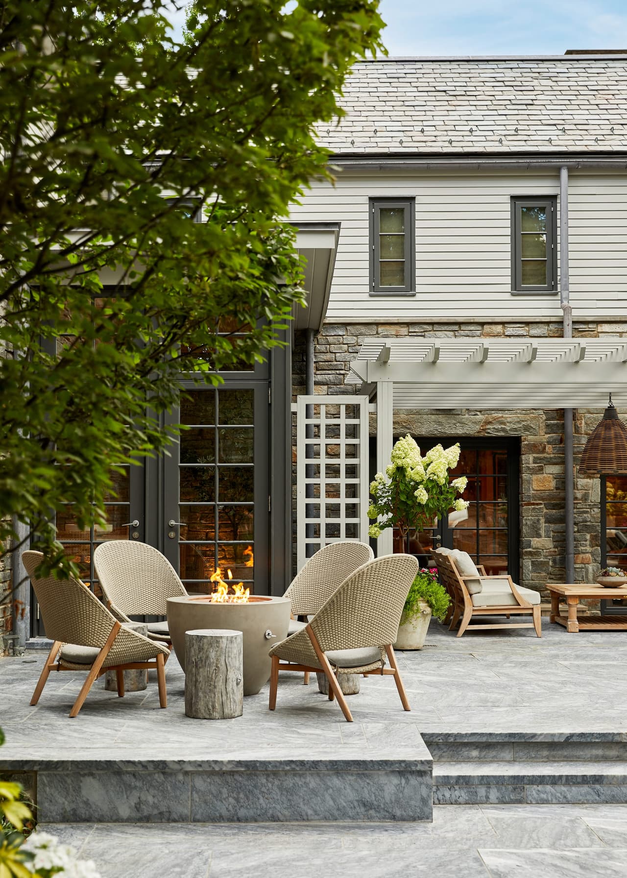 Woven chairs around a round fire table on a stone terrace, against the shingles and lattice screen of a house