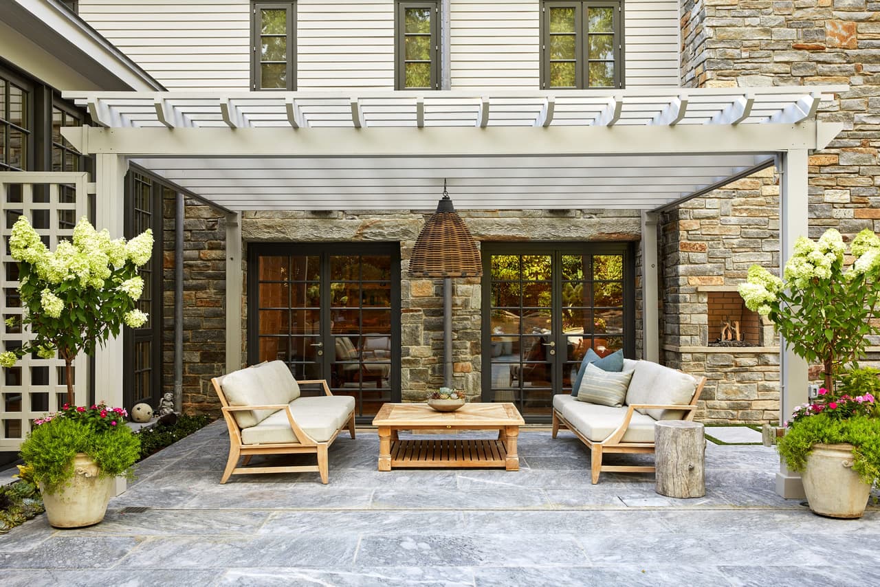 Teak lounge seating under a white pergola, against a stone wall with glass doors behind