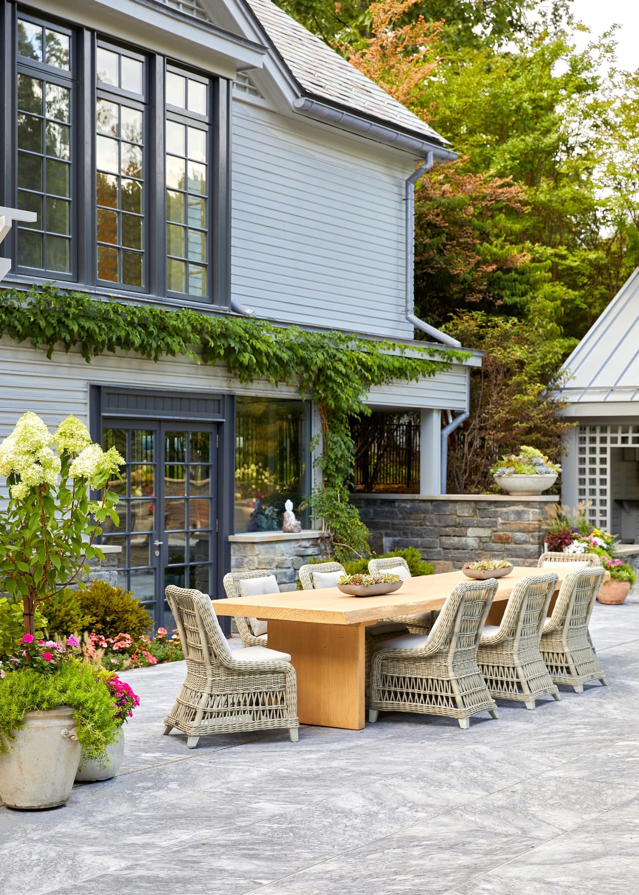 Outdoor dining table with wicker chairs on a patio beside the house, with hydrangeas in planters