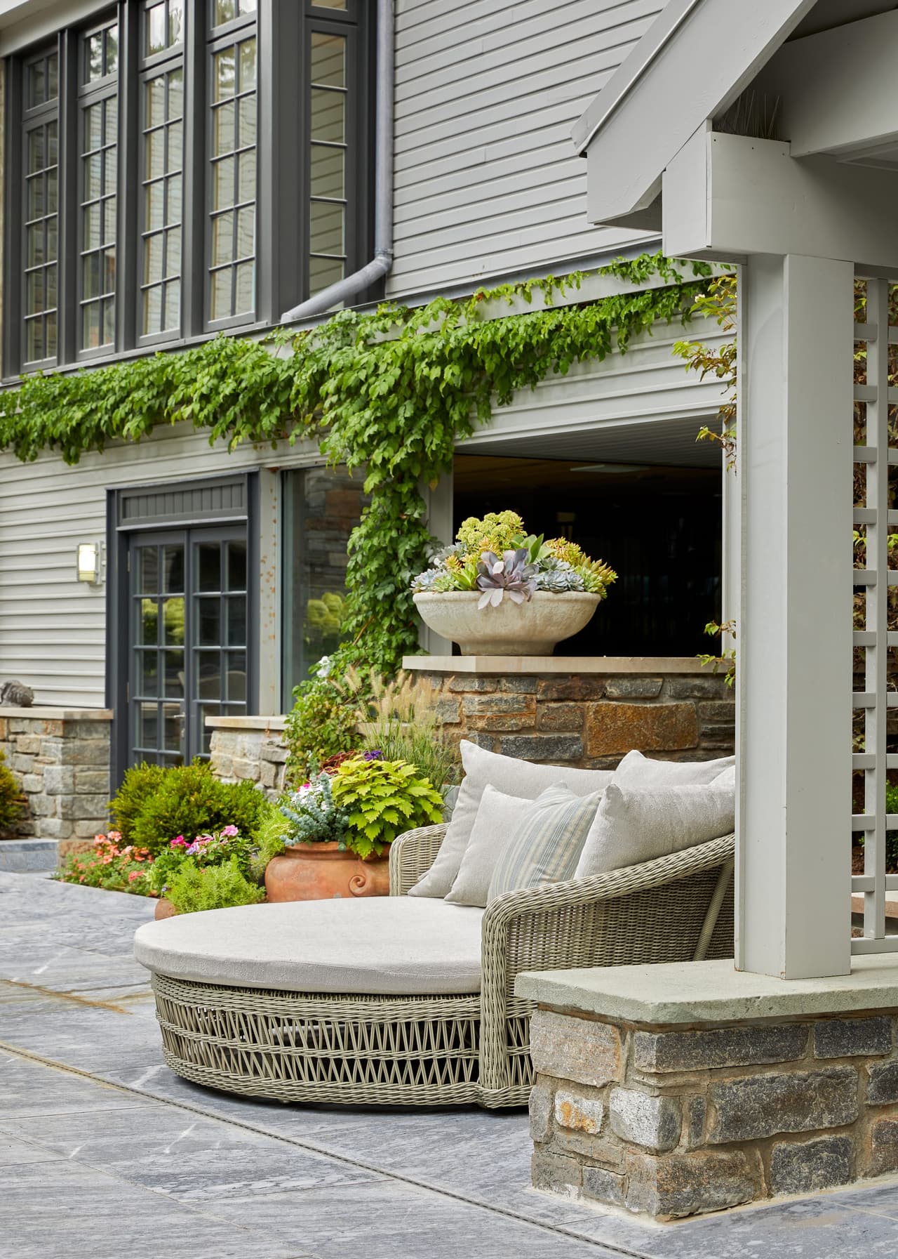 Curved outdoor daybed on a stone terrace below a vine-covered wall, beside a stone planter and a window