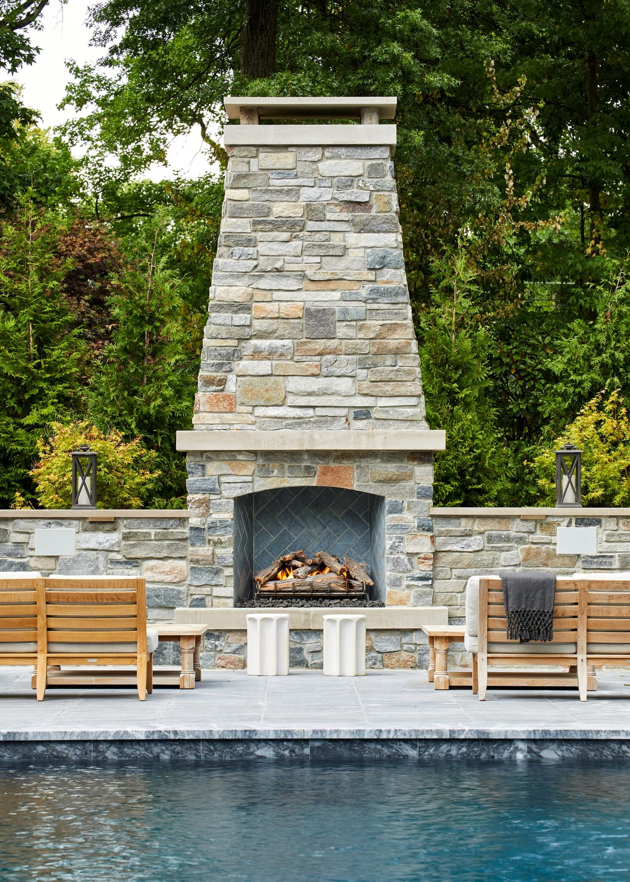 Stone outdoor fireplace rising above the pool terrace of a Westfield, NJ backyard, with teak benches either side of the firebox