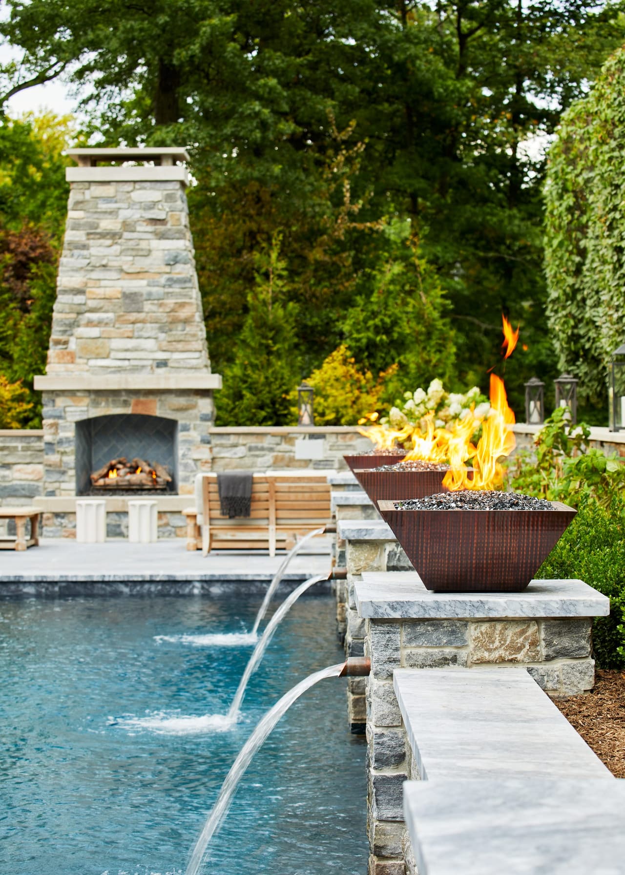 Pool with a tall stone outdoor fireplace, fire bowls on stone piers and water spouts arcing into the water
