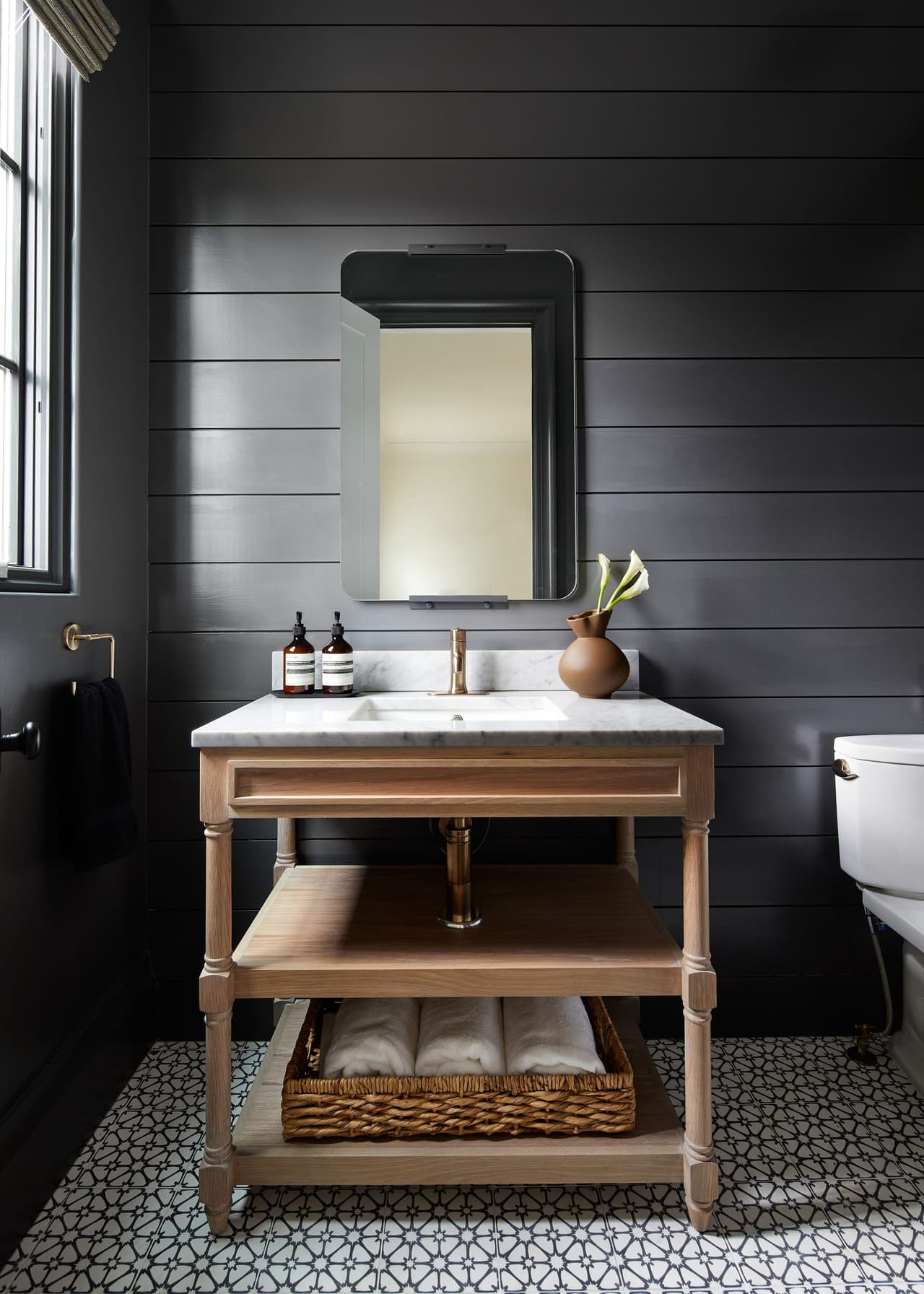 Powder room with black shiplap walls, a wooden washstand with a marble top, brass fittings and a patterned tile floor
