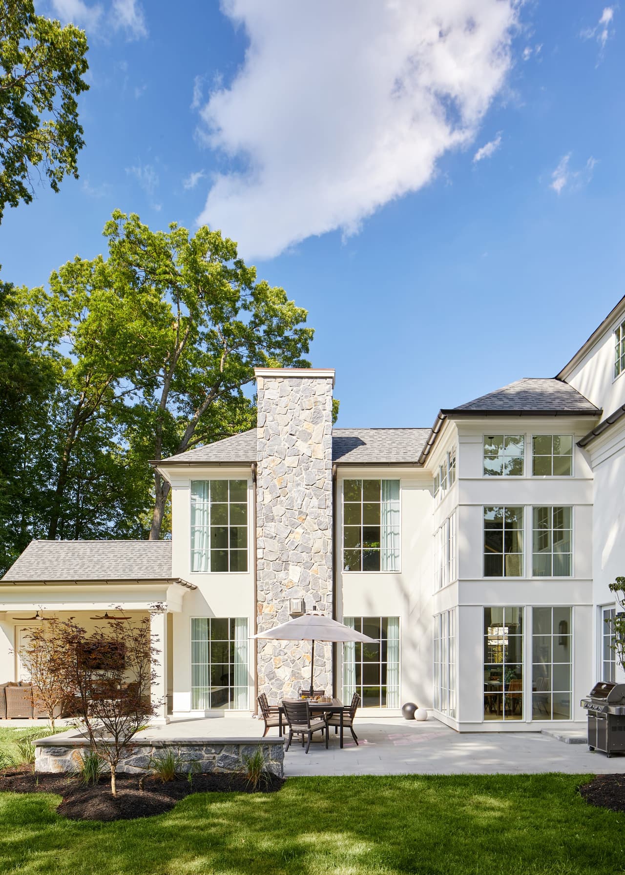 Back of a shingle and stone Westfield, NJ home renovation across the lawn, with two storeys of windows, a stone chimney and an umbrella on the terrace