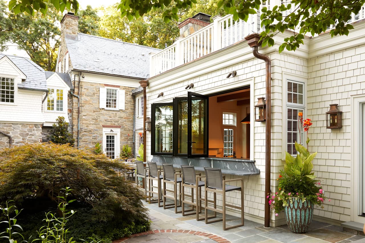 Outdoor bar counter and stools along the shingle and stone rear wall of a Westfield, NJ home renovation, with the pass-through window folded open above it
