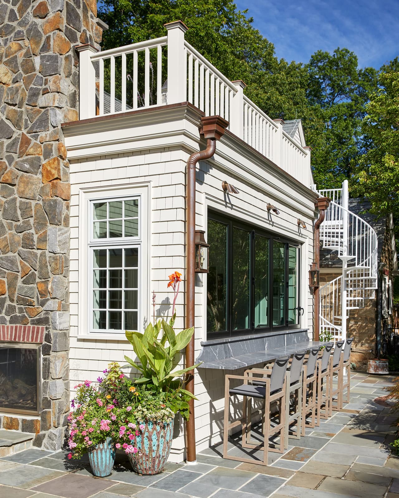 Outdoor bar and stools under the pass-through windows of a Westfield, NJ renovation, below a balustraded roof terrace with a copper leader on the stone wall