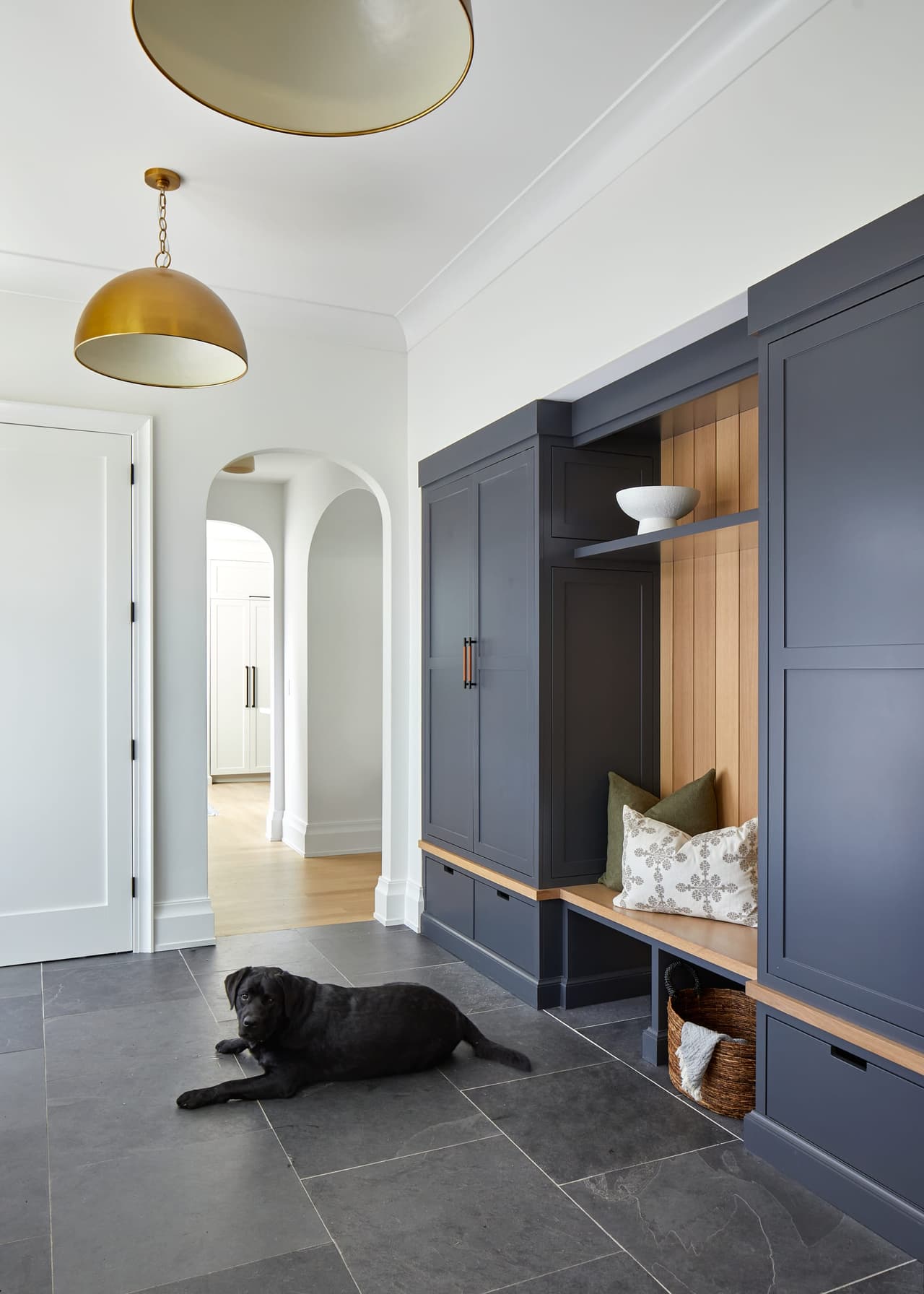 Black dog lying on the stone floor of a Westfield, NJ mudroom renovation, below charcoal cabinetry and a bench, with an arched opening to the hall beyond