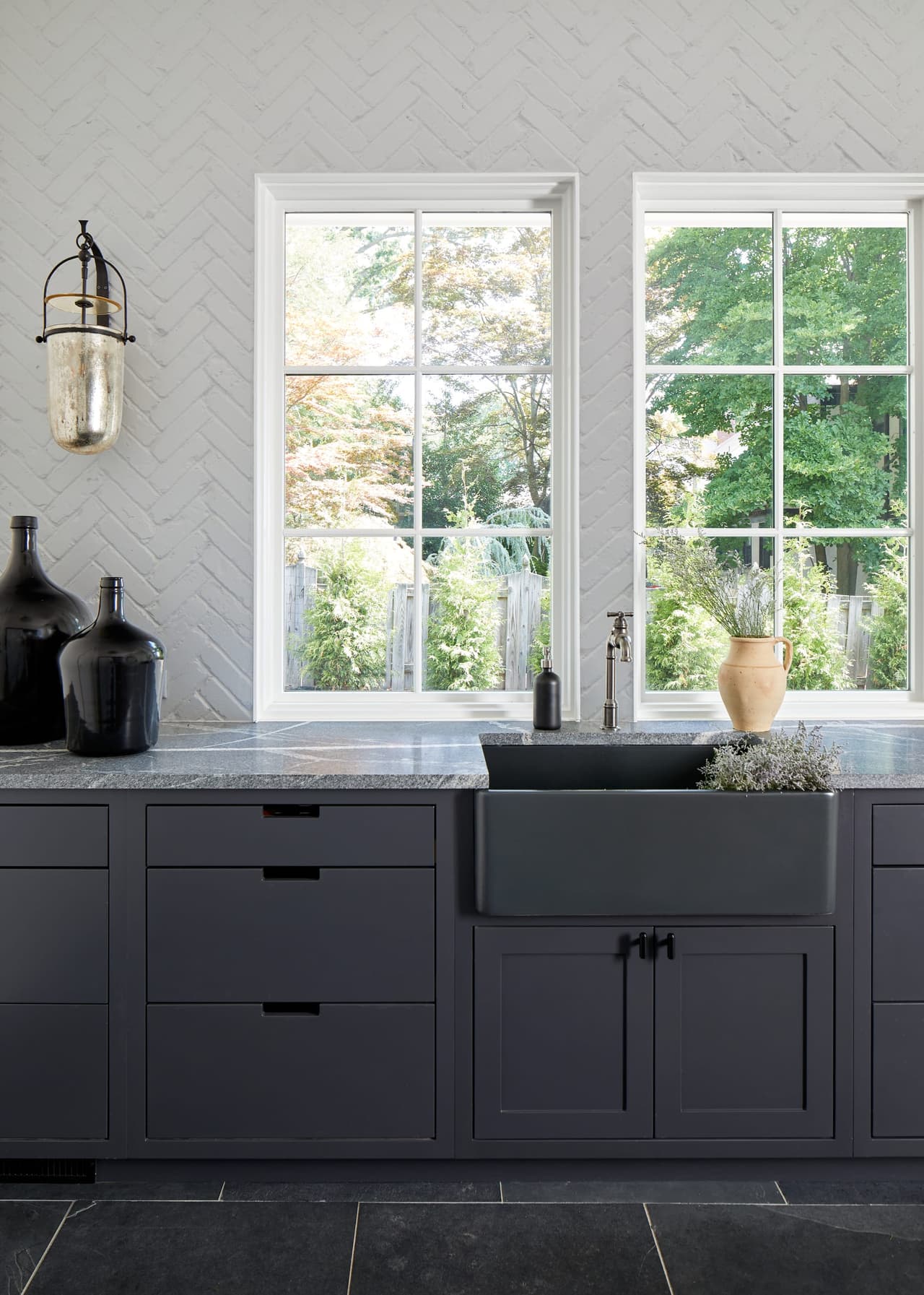 Black apron sink in navy base cabinets between two windows in a Westfield, NJ mudroom renovation, against a pale brick wall