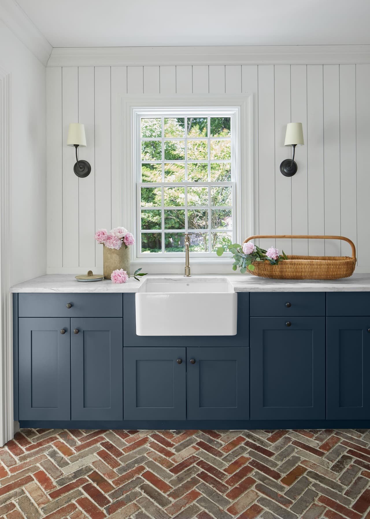 Apron-front sink in navy base cabinets under a window, with sconces either side and a herringbone brick floor, in a Westfield, NJ renovation