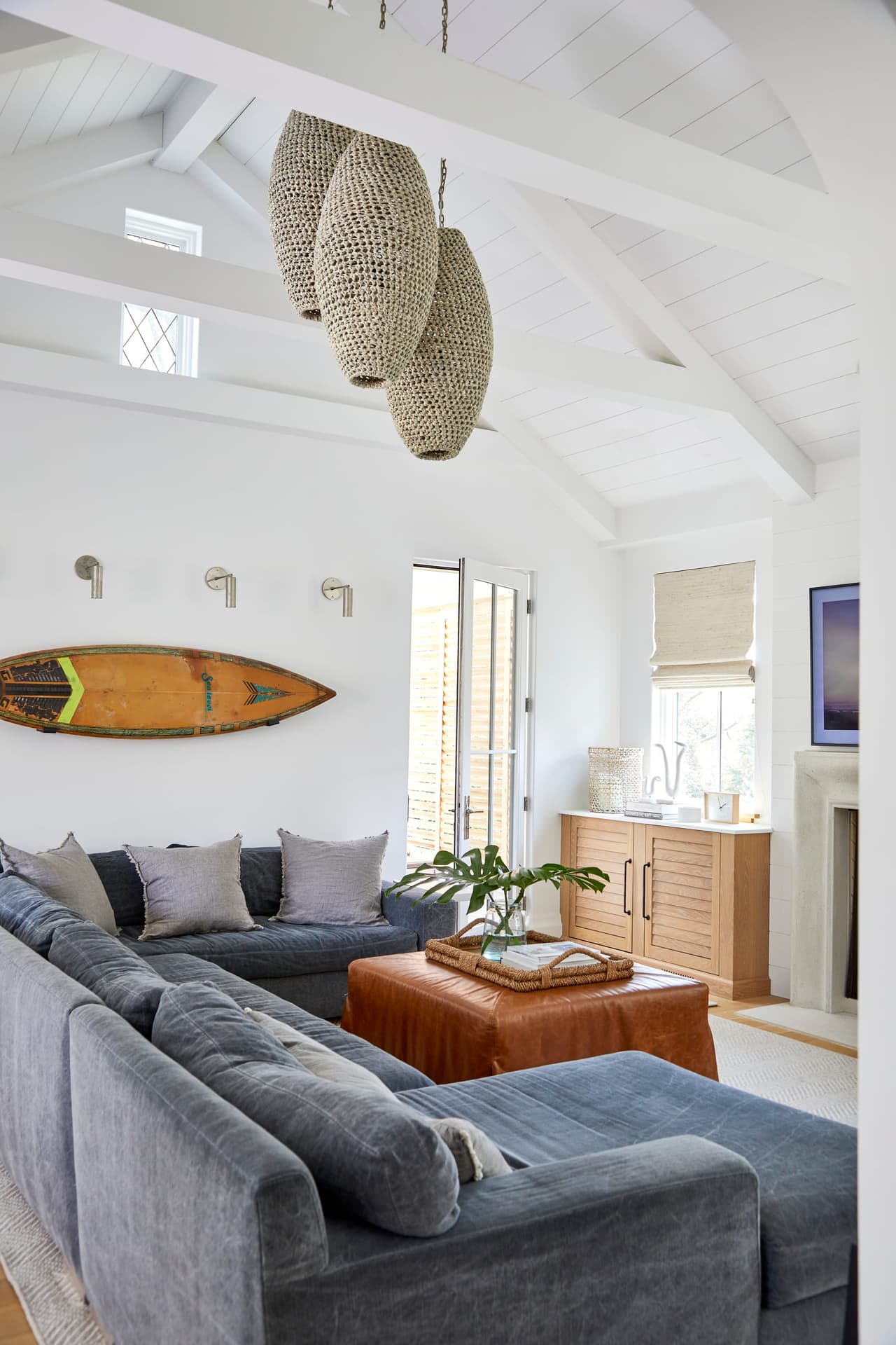 Family room of a Westfield, NJ new home under a vaulted ceiling, with woven pendants, a gray sectional and a surfboard leaning against the wall
