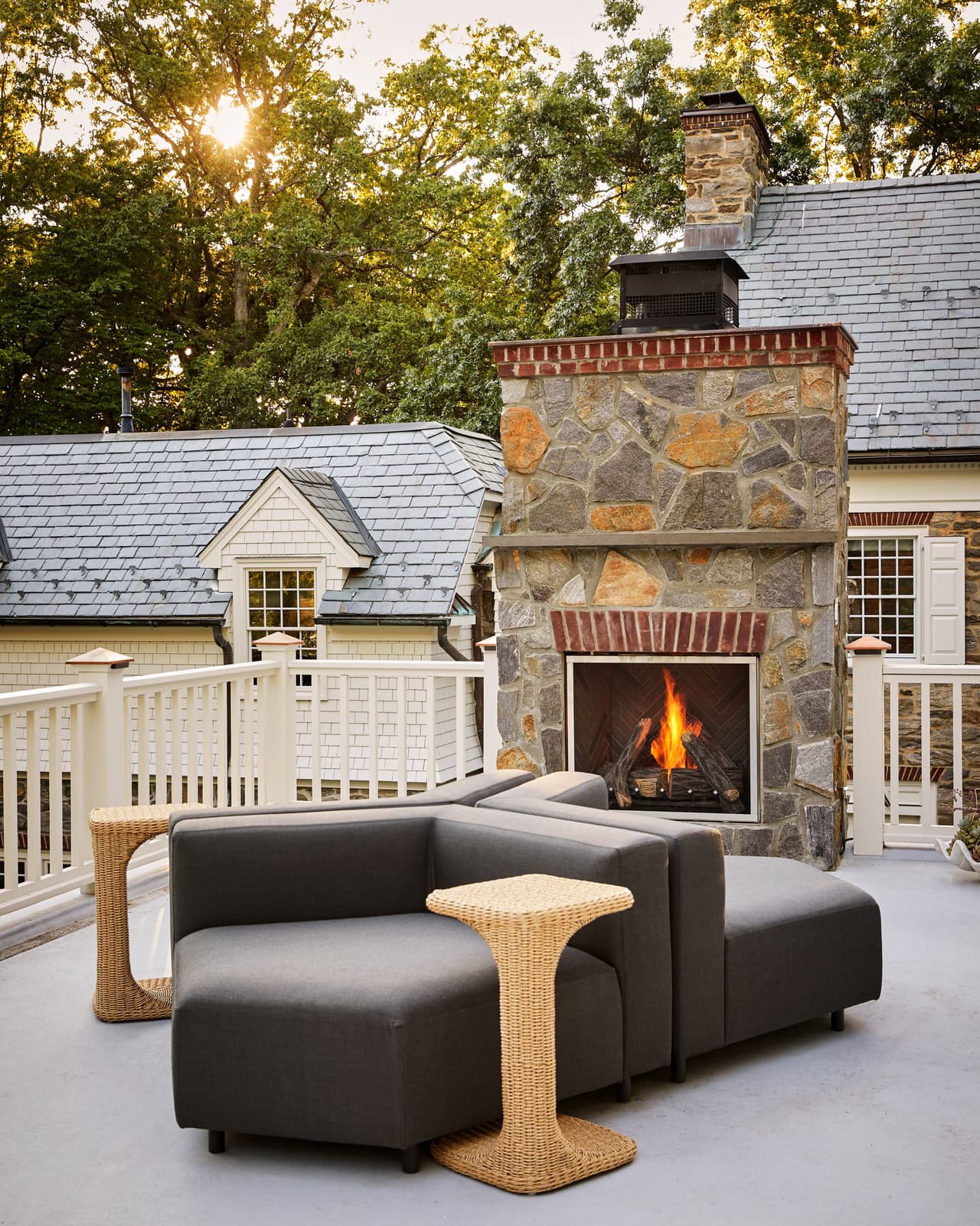 Rooftop terrace at a Westfield, NJ stone and shingle renovation, with a lit stone fireplace, a railing in white balusters and the sun through the trees, built by Michael Robert Construction