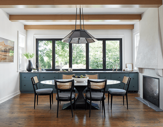 Dining room with black-framed windows, a round table with cane-backed chairs, a dark dome pendant and a teal built-in sideboard