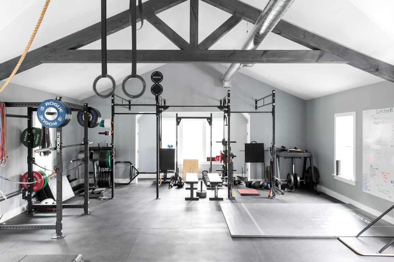 Home gym under exposed timber trusses, with a squat rack, rings, a sled track and rubber flooring the length of the room