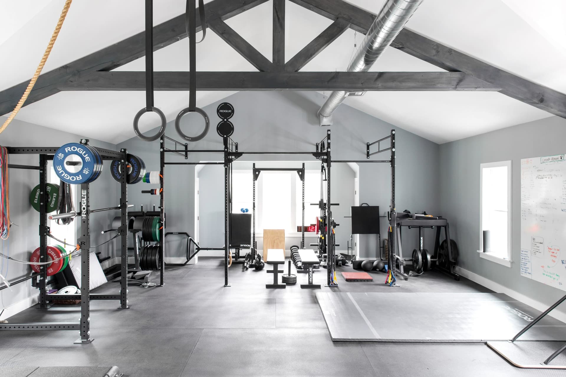Home gym under exposed timber trusses, with a squat rack, rings, a sled track and rubber flooring the length of the room