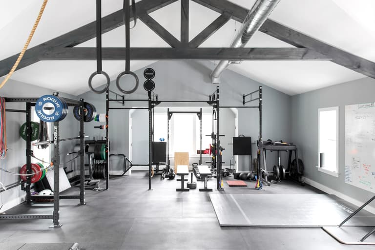 Home gym under exposed timber trusses, with a squat rack, rings, a sled track and rubber flooring the length of the room