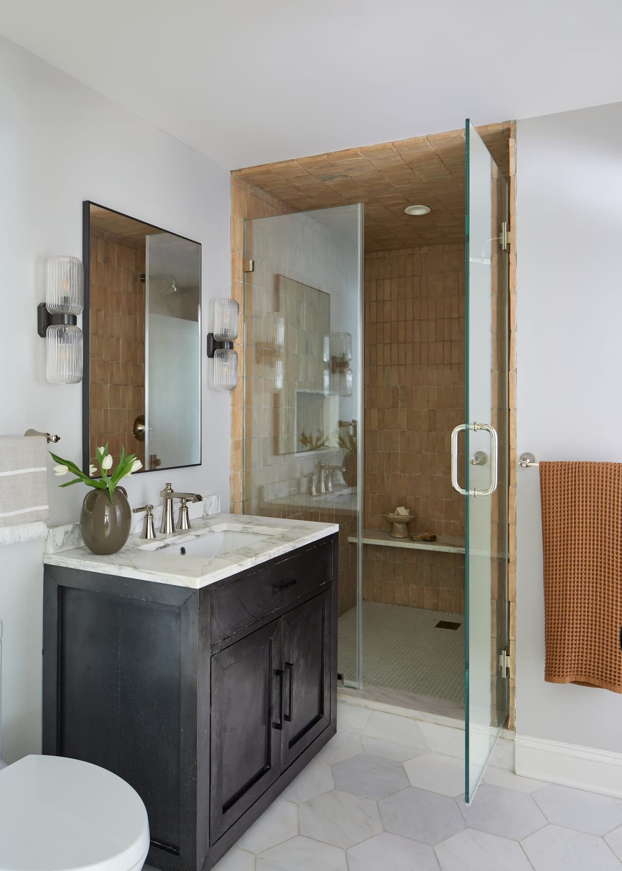 Dark stained vanity under a framed mirror between two sconces in a Westfield, NJ renovation, beside a glass shower on a hexagonal tile floor