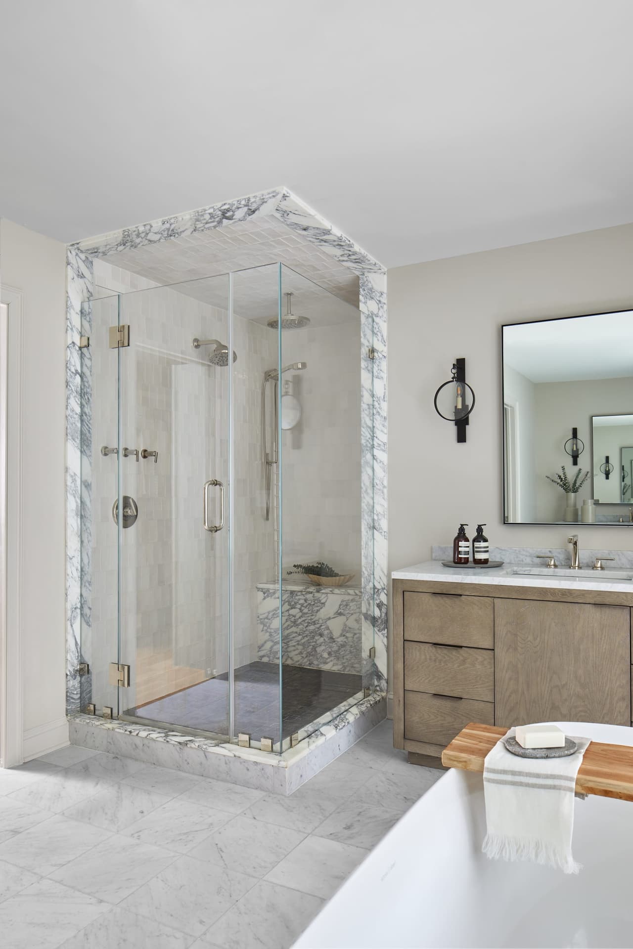 Primary bathroom of a Westfield, NJ renovation with a marble-lined corner shower in clear glass, an oak vanity and a tub in the foreground
