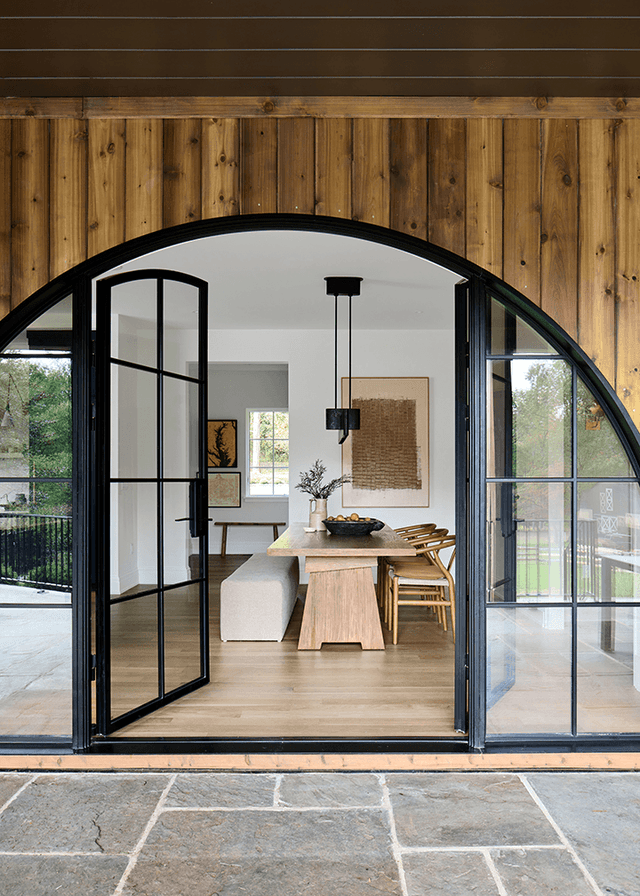 Arched black steel and glass door opening from the stone terrace into the dining room of a Mendham, NJ home, under a reclaimed wood wall