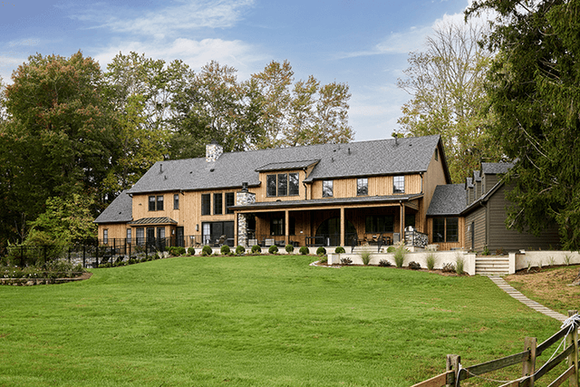 Back of a cedar and stone Mendham, NJ home across the lawn, with a long covered porch, stone chimneys and a split-rail fence along the field