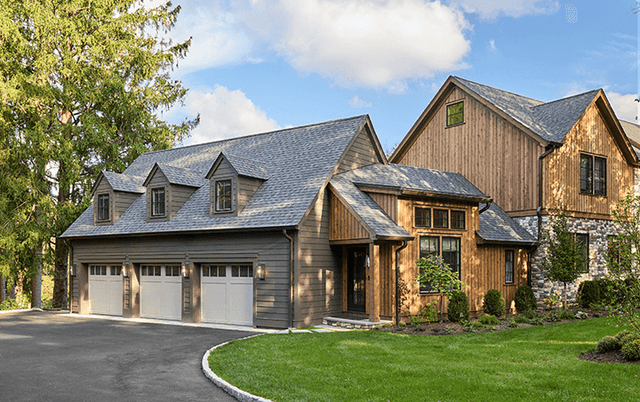 Three-bay garage wing of a cedar and stone Mendham, NJ home, with gabled dormers above and a stone chimney on the main house beyond