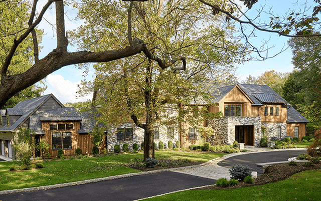 Front of a cedar and stone home in Mendham, NJ seen under a mature oak, with gabled dormers and a curving drive, built by Michael Robert Construction