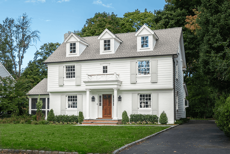 Front of a white painted brick home with a steep slate-look roof, arched dormers and clipped planting across the front