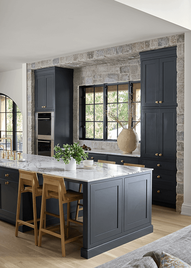 Kitchen of a Mendham, NJ home with navy custom cabinetry down both sides of an exposed stone wall and a marble-topped island with oak stools