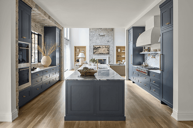 Navy kitchen of a Mendham, NJ home seen through an arched doorway, with a marble-topped island, an exposed stone wall and cabinetry down both sides