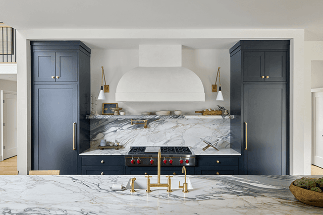 Range wall of a Mendham, NJ kitchen, with a plaster hood between navy cabinets and a marble slab carried up behind the brass-trimmed cooktop