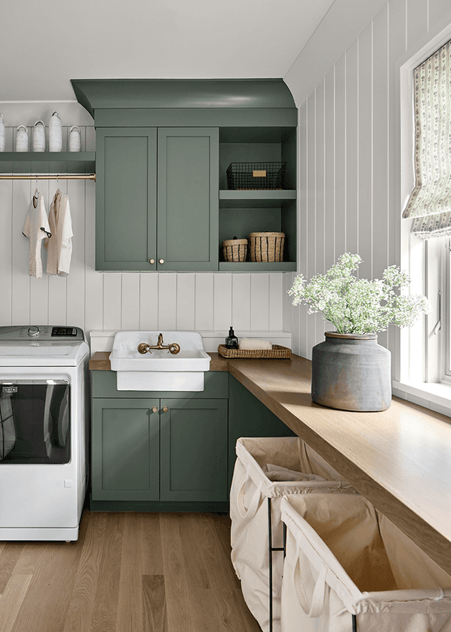 Laundry room in a Mendham, NJ home with teal custom cabinets over an apron sink, a brass tap, a vertical shiplap wall and canvas hampers below the counter