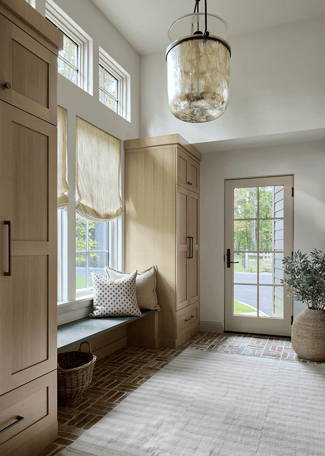 Mudroom of a Mendham, NJ home with custom white oak cabinetry, a window seat with a Roman shade and a brick floor