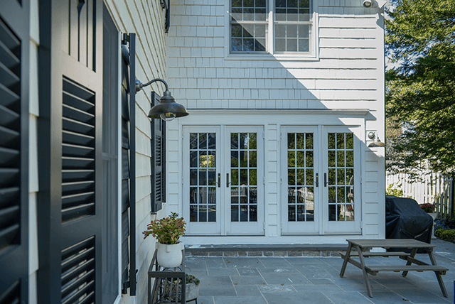 Bluestone patio of a Westfield, NJ home renovation, with a gooseneck barn light on white shingle siding, louvered shutters and French doors opening onto the terrace