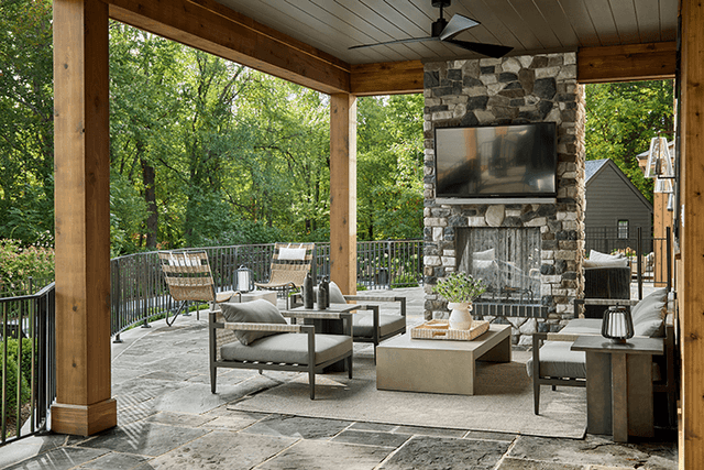 Covered patio at a Mendham, NJ home, with a stone fireplace and television under a timber ceiling, and an outdoor kitchen along the far wall