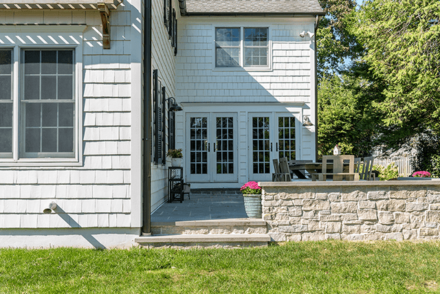 Raised stone patio wall against the blue-gray shingles of a Westfield, NJ exterior renovation, with a pergola over the terrace beyond
