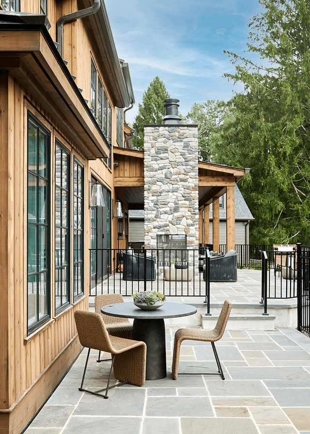 Bluestone patio along the cedar-clad rear elevation of a Mendham, NJ home, with a round table and two chairs beside the stone chimney