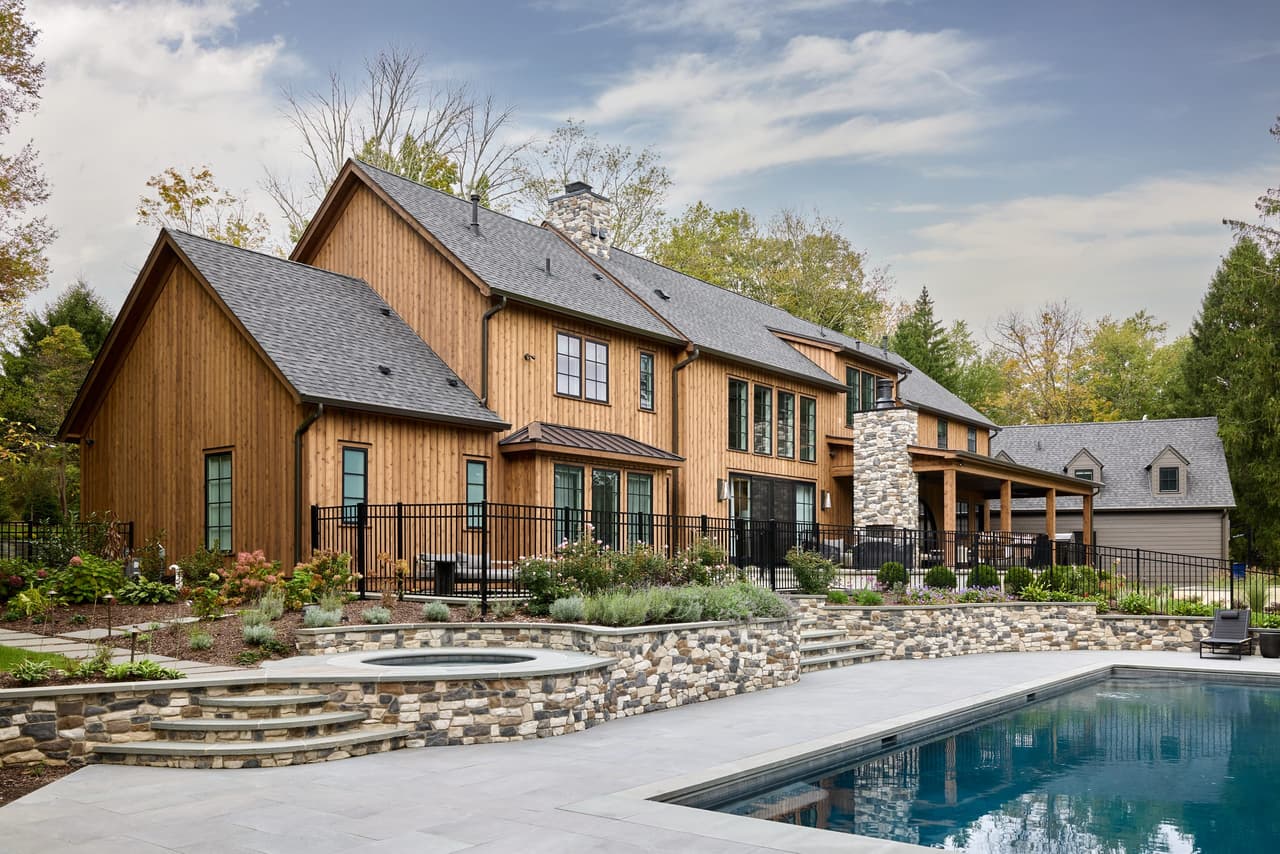 Pool and spa below the cedar and stone rear elevation of a Mendham, NJ home, with a dry-laid stone retaining wall between them