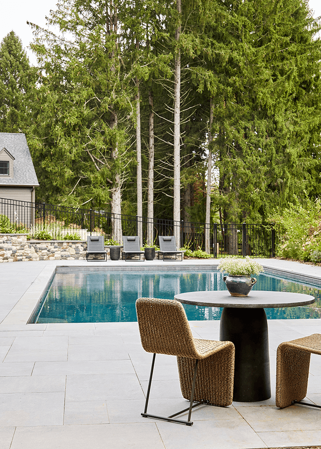 Pool and stone patio at a Mendham, NJ home, with woven chairs around a round table, loungers along the far side and pines behind