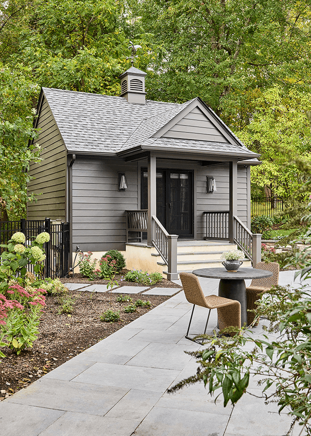 Shingled poolhouse at a Mendham, NJ home, with a cupola, a black door up two steps and a bluestone path through the planting